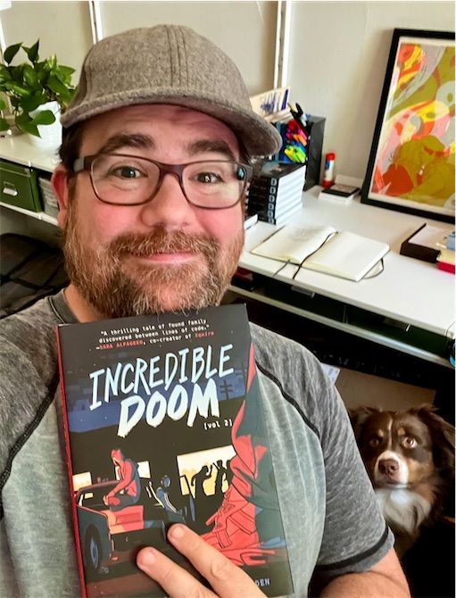 Matt holding up a copy of Incredible Doom Vol. 2 and smileing, while a dog looks on in the background. 