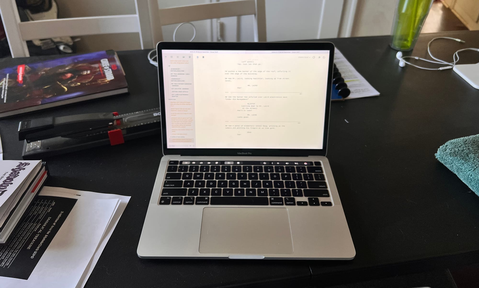 An open laptop displaying a script sits on a dark table surrounded by books, papers, a ruler, and a water bottle, suggesting focused writing work.