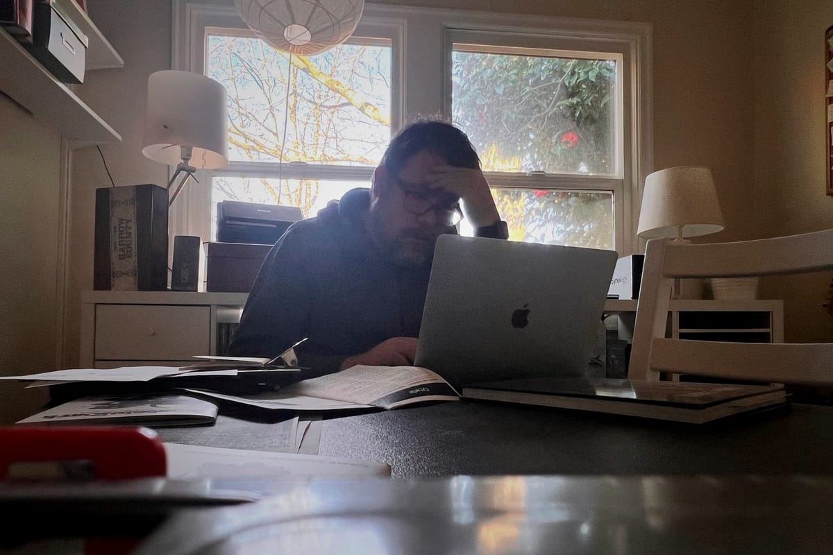A person sits at a cluttered table, leaning over a laptop with a hand on their forehead. Papers and books surround them as daylight streams through windows behind.